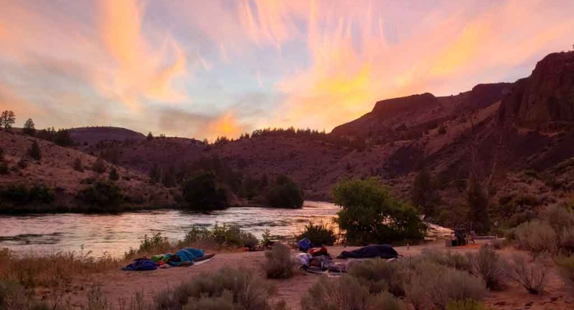 the sun rises behind a ridge in the background of a campsite along a river on an outward bound trip